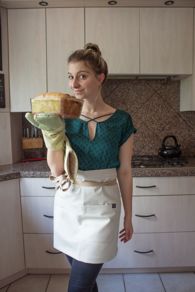 A girl in an apron holding a baked loaf of bread and smirking at the camera.
